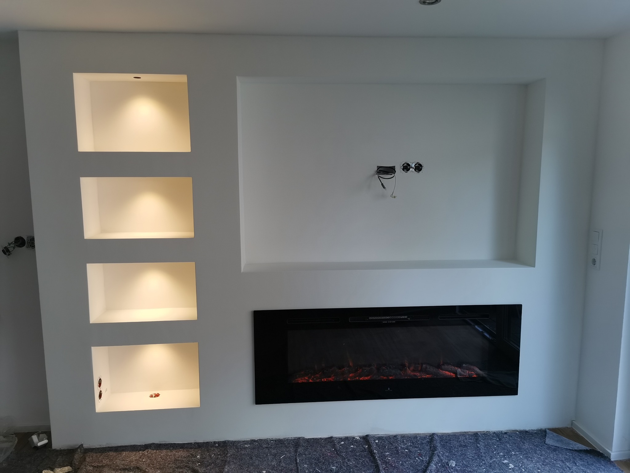 Biała ściana z wnękami oświetlonymi punktowo i elektrycznym kominkiem. Widoczne gniazdka elektryczne. Podłoga zabezpieczona folią malarską.