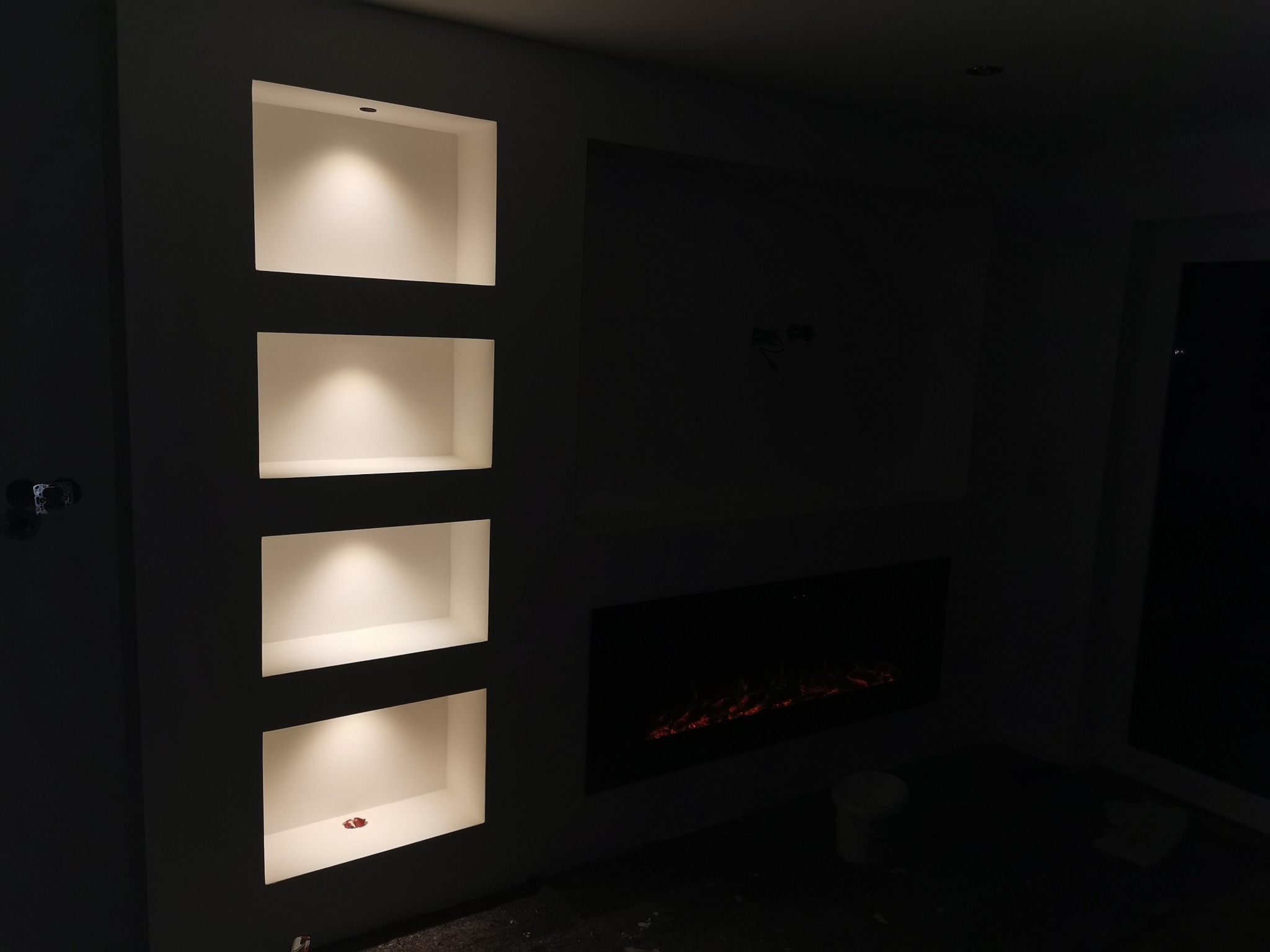 Nowoczesne wykończenie wnętrza: ściana z wnękami oświetlonymi punktowo i elektrycznym kominkiem. Minimalistyczny design, ciemne tło, subtelne światło.