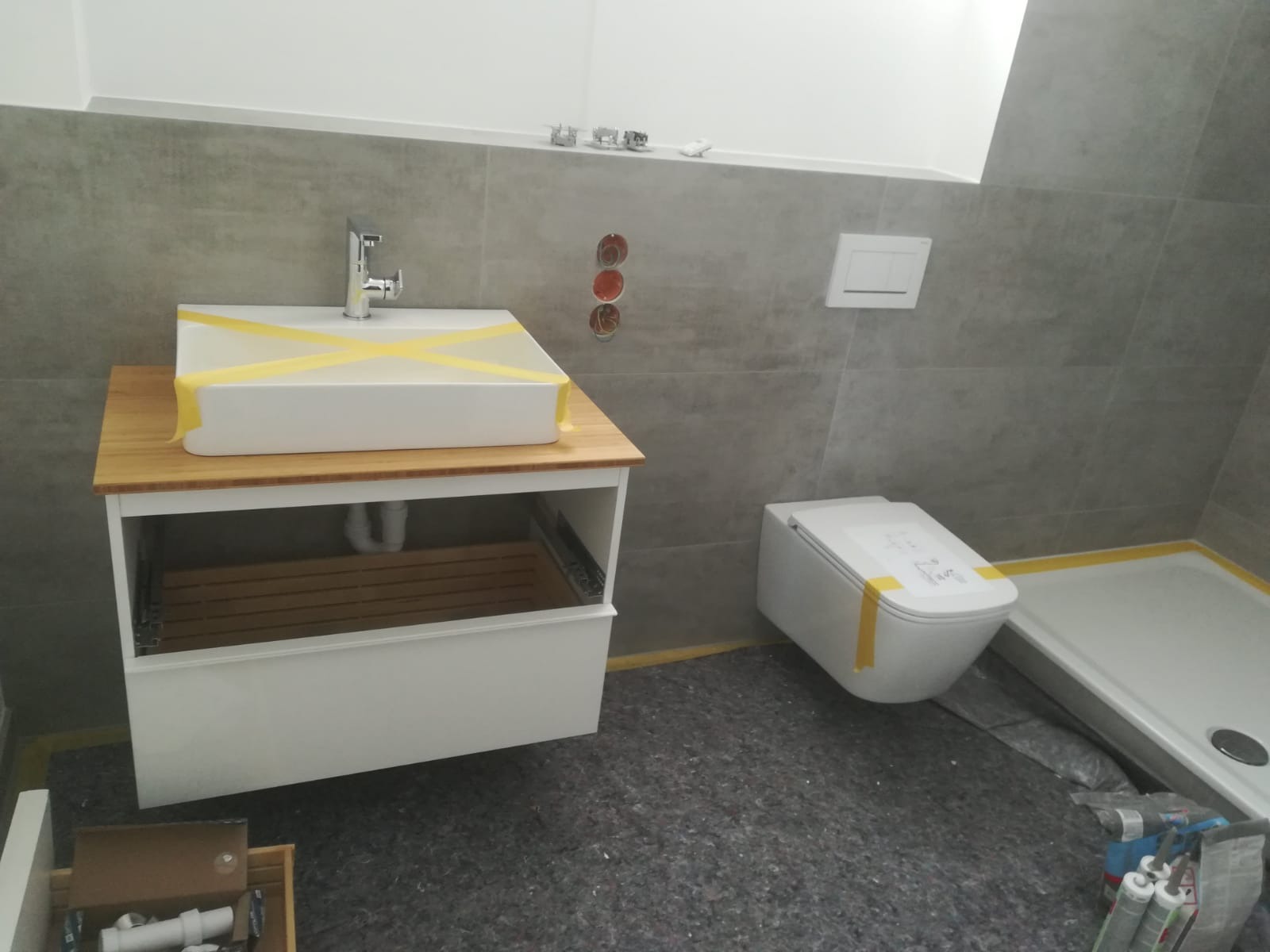 Nowoczesna łazienka w trakcie wykończenia: biała umywalka na drewnianej szafce, podwieszana toaleta i brodzik. Szare płytki na ścianach, zabezpieczenia taśmą malarską.