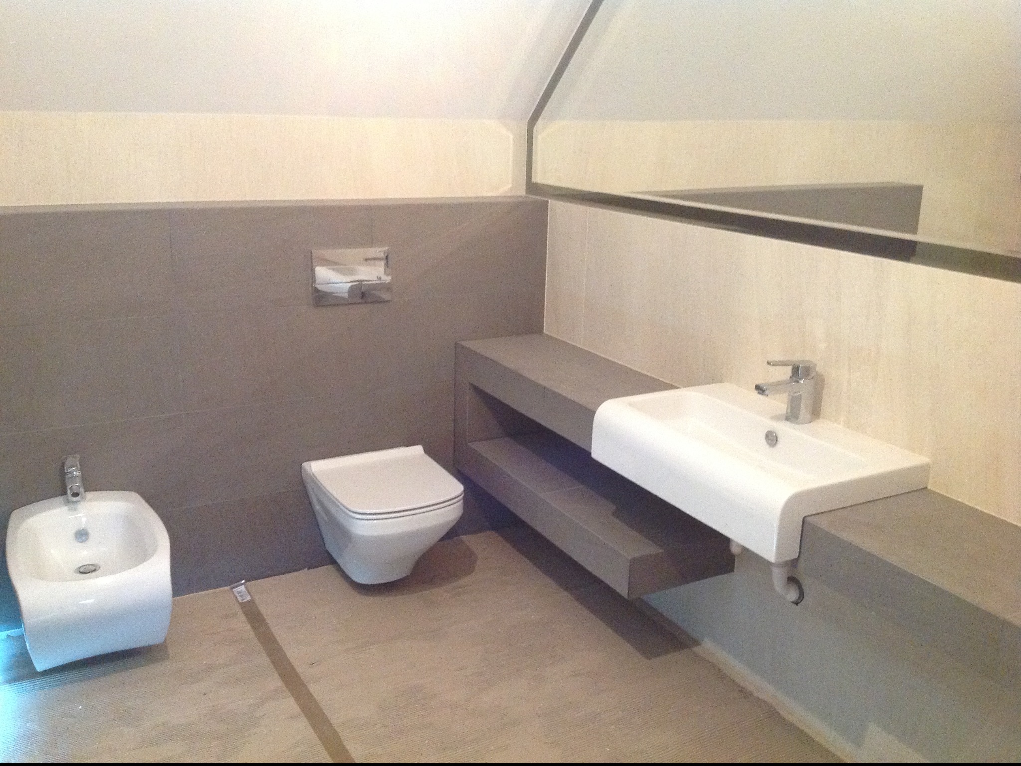 Nowoczesna łazienka na poddaszu z białym bidetem, podwieszaną toaletą i umywalką na szarym blacie. Ukośny sufit i duże lustro dodają przestrzeni.