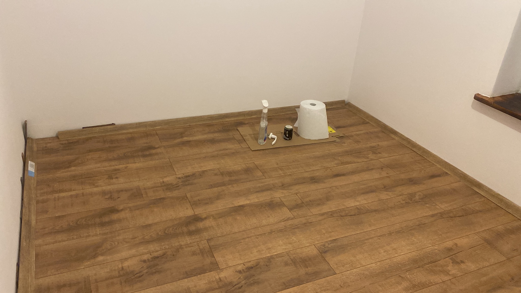 Ukończona podłoga z paneli w rogu pokoju, z widocznymi akcesoriami do czyszczenia na kartonie. Ciepłe odcienie brązu i beżu, naturalne światło.