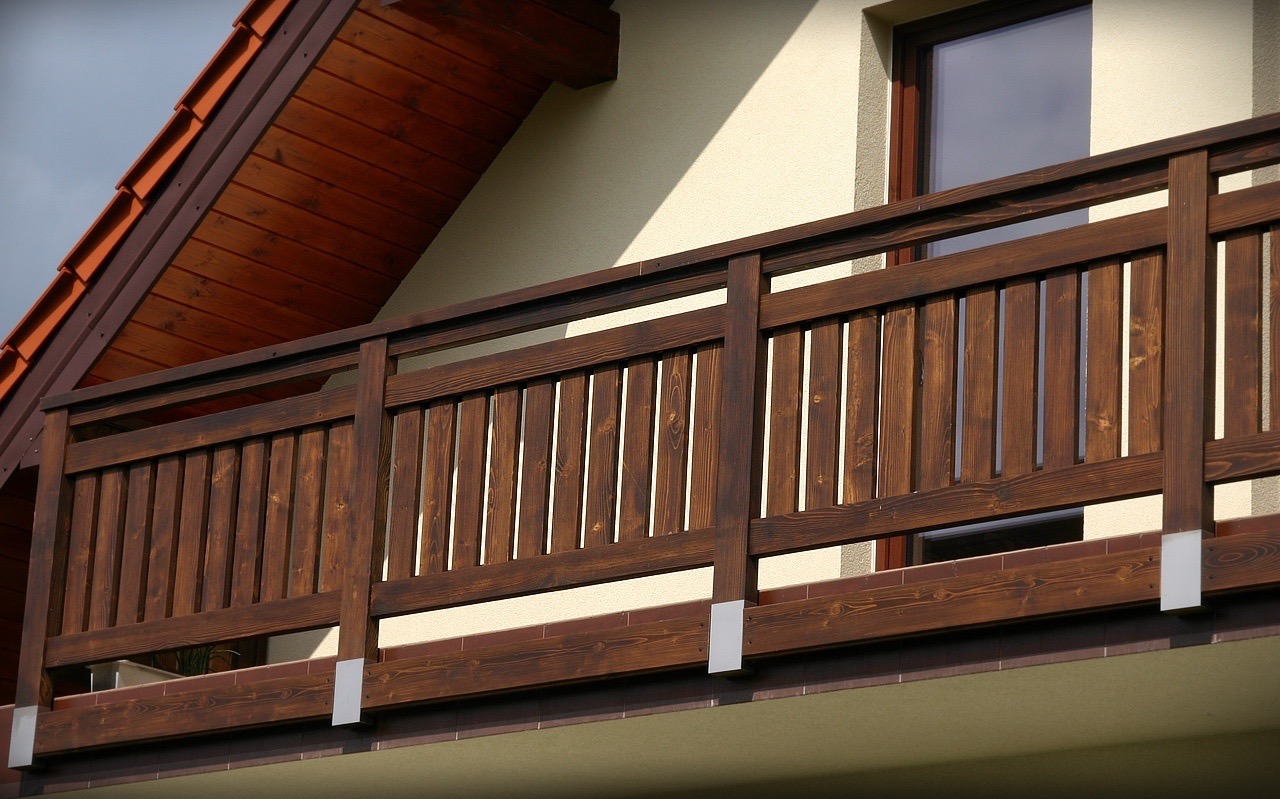 Drewniana balustrada balkonu z ciemnego drewna, widok z dołu, fragment dachu z czerwoną dachówką, okno w tle, jasna elewacja budynku.