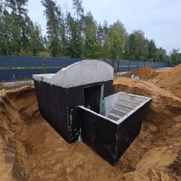 Krzysztof Ka - Betonowy schron/piwnica w ziemi z wejściem schodami, zabezpieczony czarną izolacją, na tle zieleni i ogrodzenia panelowego. Widok z góry.