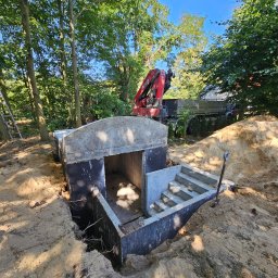 Krzysztof Ka - Montaż betonowego szamba z wejściem schodkowym w ziemi, widoczny dźwig i wykop. Praca w toku, teren zalesiony, słoneczny dzień.