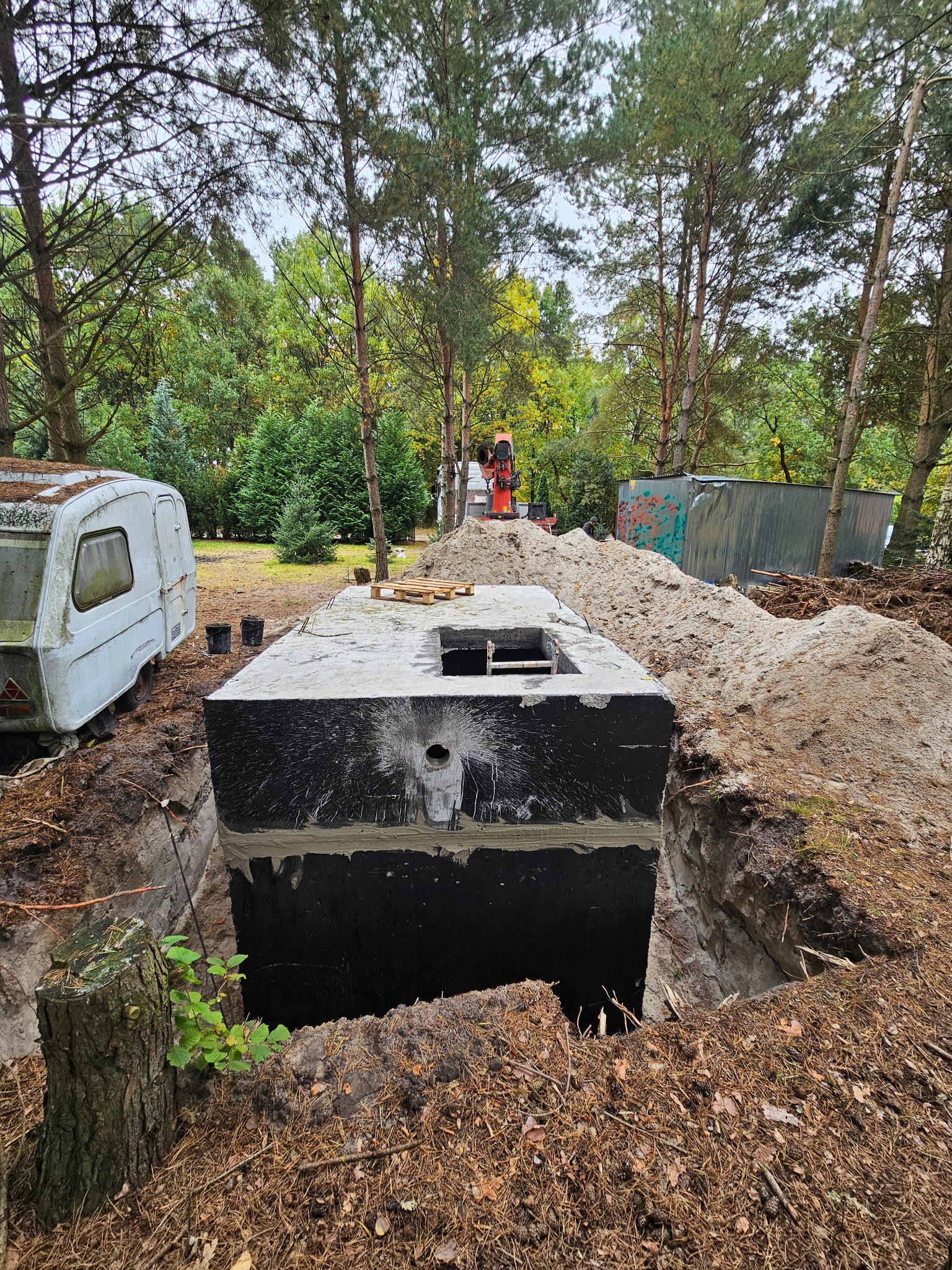 Ciemny, betonowy szambon na wykopie, otoczony ziemią i drzewami. Widoczny fragment przyczepy kempingowej i sprzęt budowlany w tle. Ujęcie z bliska, podkreślające detale konstrukcji.