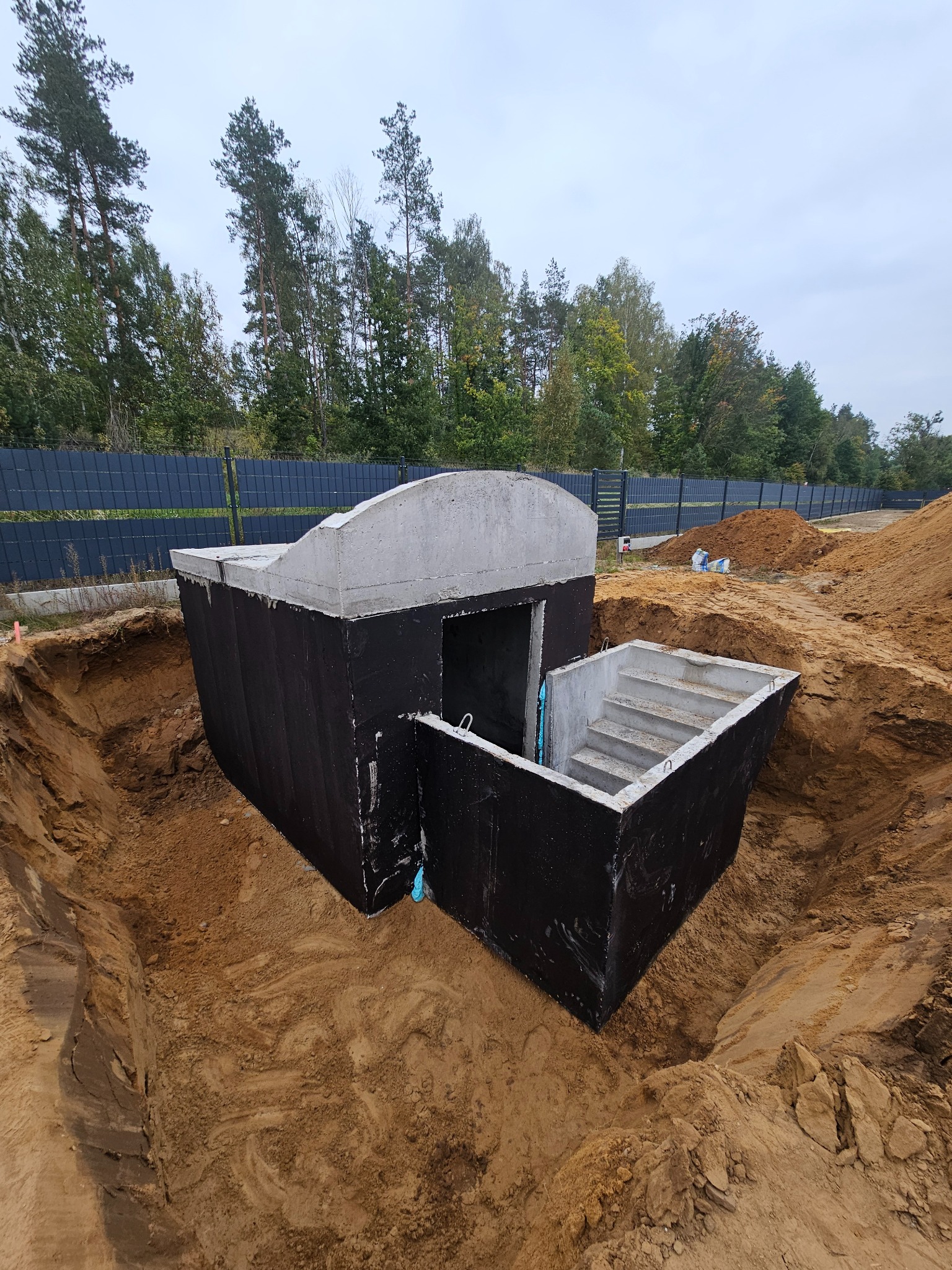 Betonowy schron/piwnica w ziemi z wejściem schodami, zabezpieczony czarną izolacją, na tle zieleni i ogrodzenia panelowego. Widok z góry.