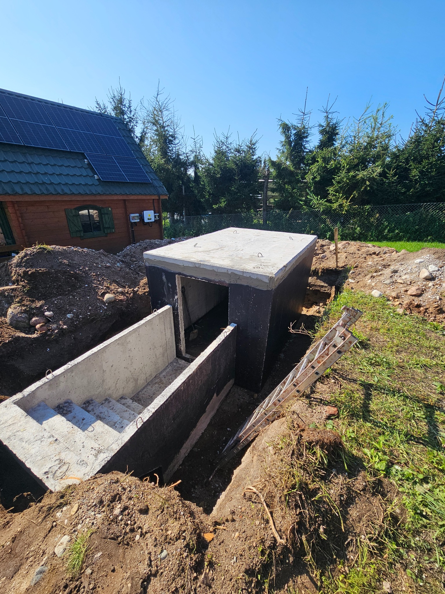 Montaż betonowego szamba z zejściem schodami w ziemi, widoczne zabezpieczenie ścian, drabina oparta o konstrukcję, dom z panelami słonecznymi w tle.