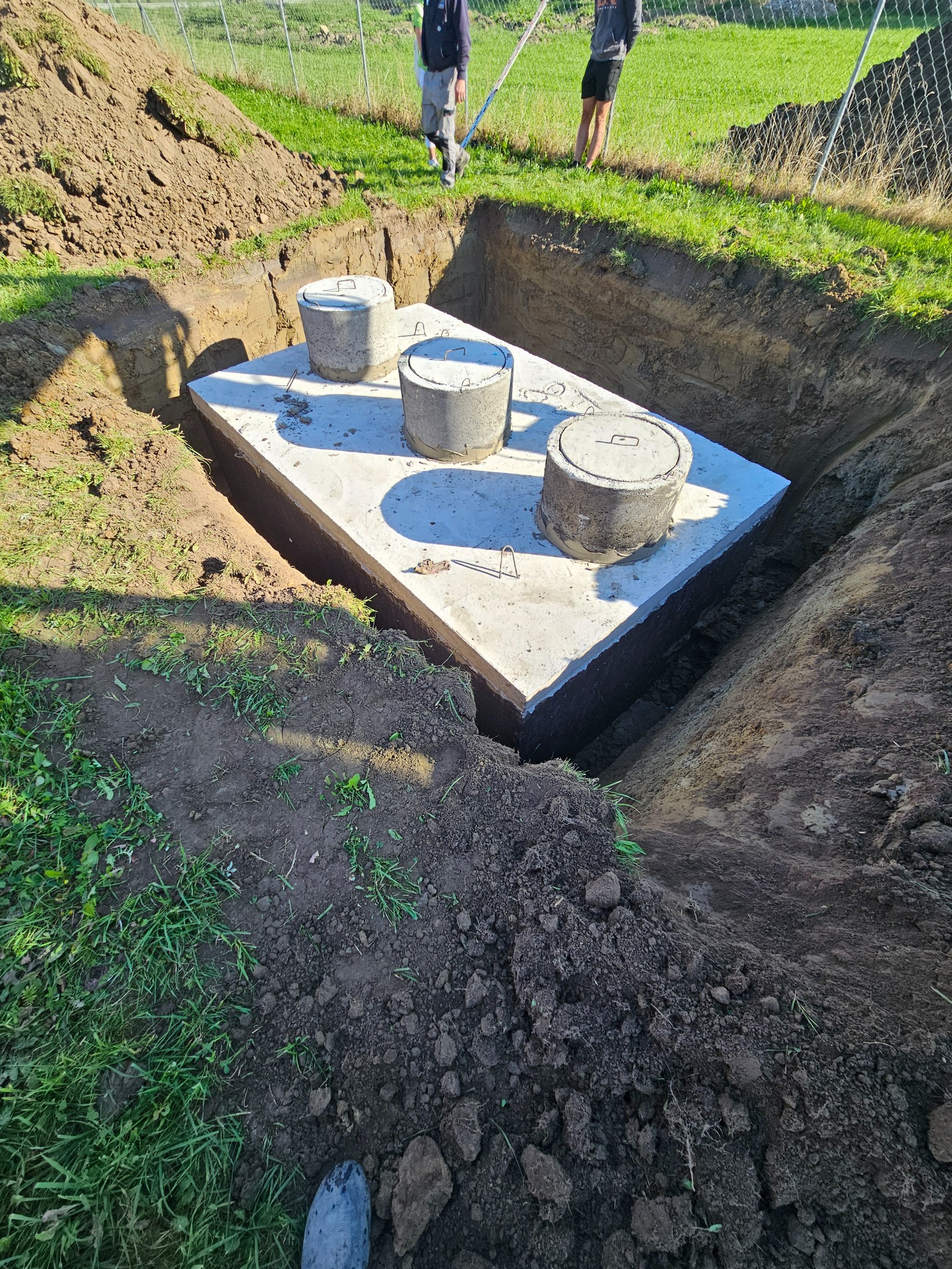 Betonowy szambo w wykopie, trzy włazy z metalowymi uchwytami, ziemia i trawa wokół, dwie osoby w tle. Widok z góry na częściowo zakopany zbiornik.
