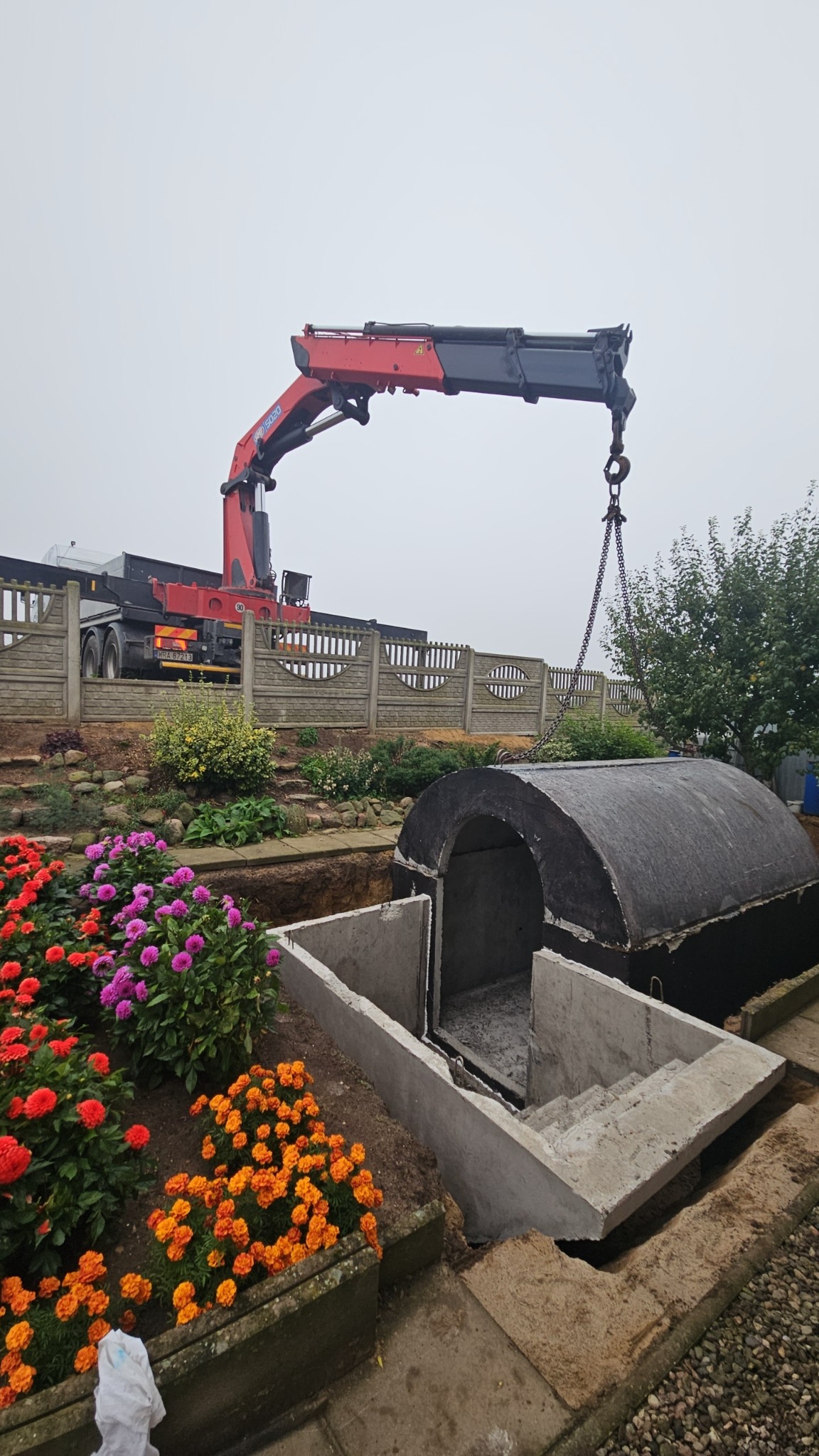 Montaż betonowego prefabrykatu z wejściem schodkowym za pomocą dźwigu na działce z kwiatami i ogrodzeniem. Etap instalacji.