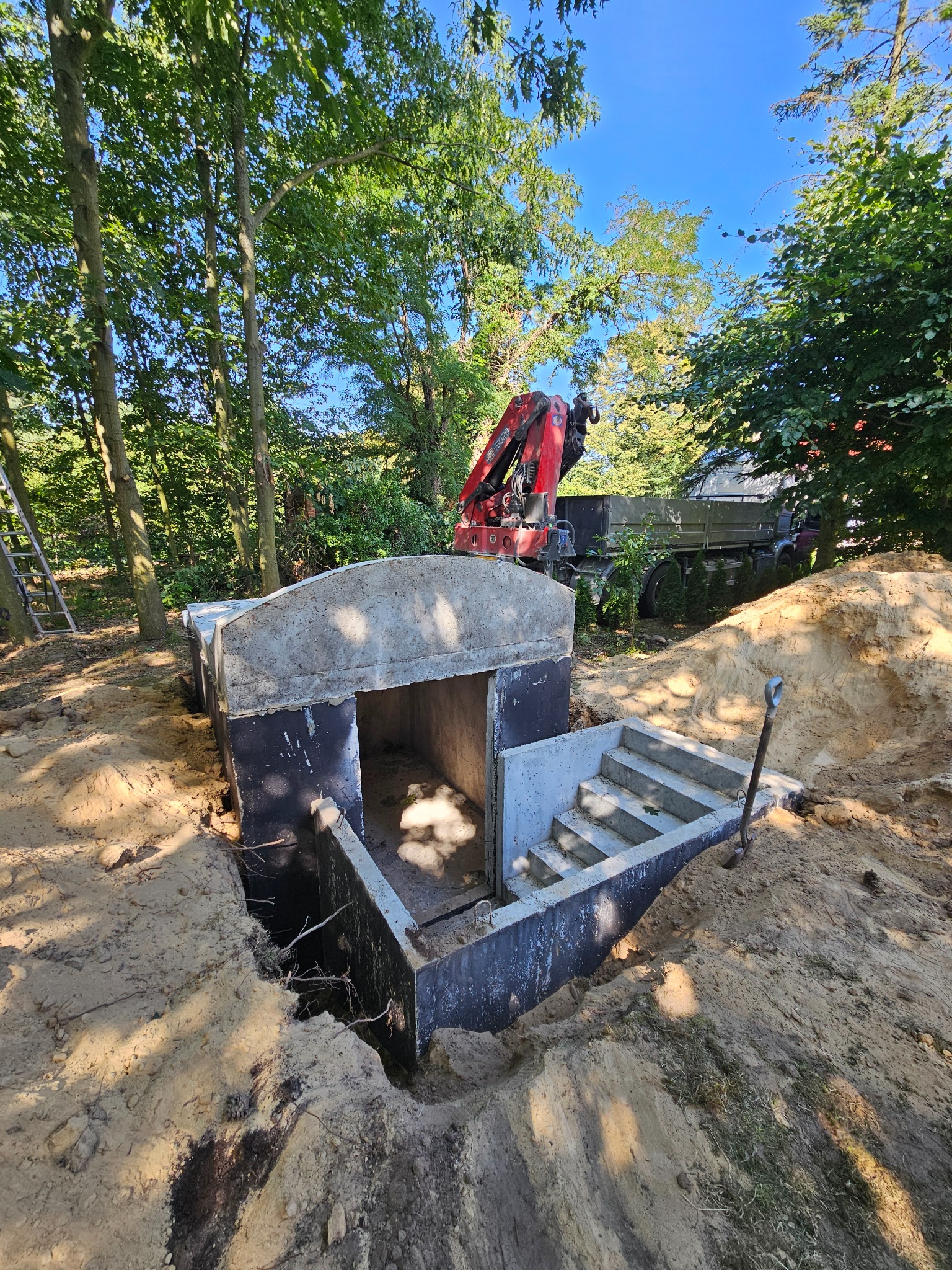 Montaż betonowego szamba z wejściem schodkowym w ziemi, widoczny dźwig i wykop. Praca w toku, teren zalesiony, słoneczny dzień.