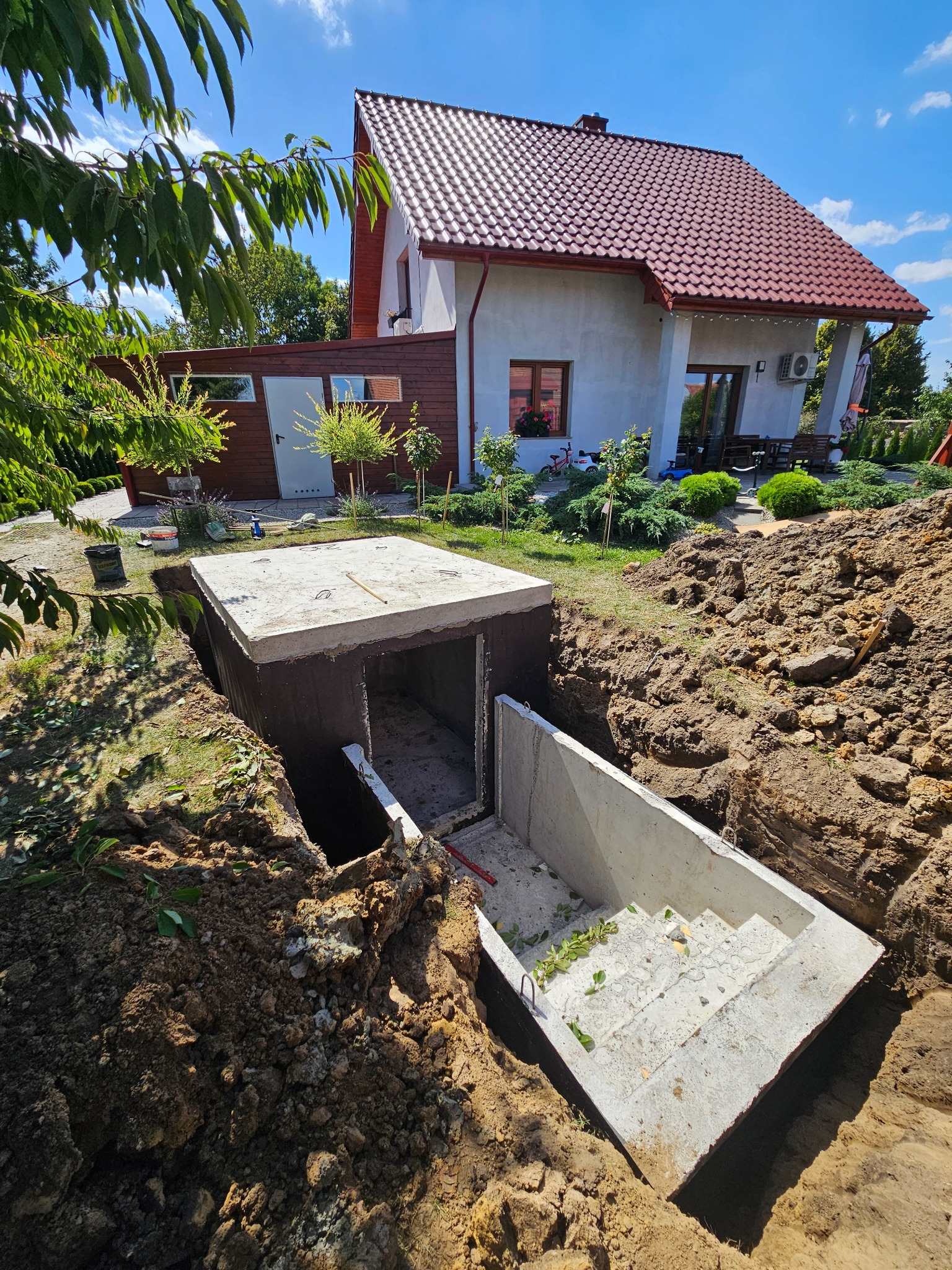 Montaż betonowego szamba z prefabrykatów na działce, widok z góry na wykop z elementami konstrukcyjnymi. W tle dom jednorodzinny i zieleń.