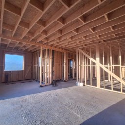 DREWNIAK HOLY WOOD GARDEN BARTOSZ KACPRZAK - Wnętrze domu w trakcie budowy, widoczne drewniane ramy ścian i stropu, surowy beton na podłodze, okno z niebieskim tłem. Etap wznoszenia konstrukcji.