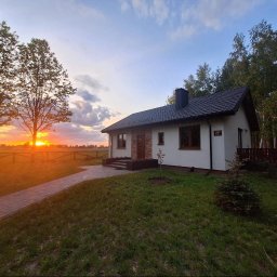 DREWNIAK HOLY WOOD GARDEN BARTOSZ KACPRZAK - Parterowy dom z ciemnym dachem i jasną elewacją o zmierzchu. Widoczny ogród z ścieżką i las w tle. Na ścianie numer 70.