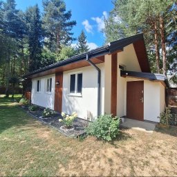 DREWNIAK HOLY WOOD GARDEN BARTOSZ KACPRZAK - Parterowy dom z białymi ścianami i drewnianymi akcentami, otoczony zielenią. Widoczny fragment trawnika i drzewa w tle. Nowoczesna architektura w leśnym otoczeniu.