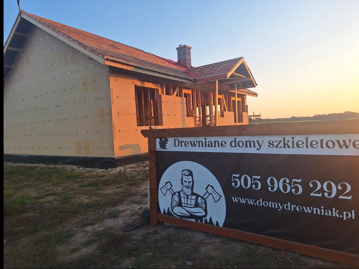 Wznoszony dom szkieletowy w ciepłym świetle zachodu słońca, z widocznym banerem reklamowym firmy 'Domy Drewniak' z numerem telefonu i adresem strony internetowej.