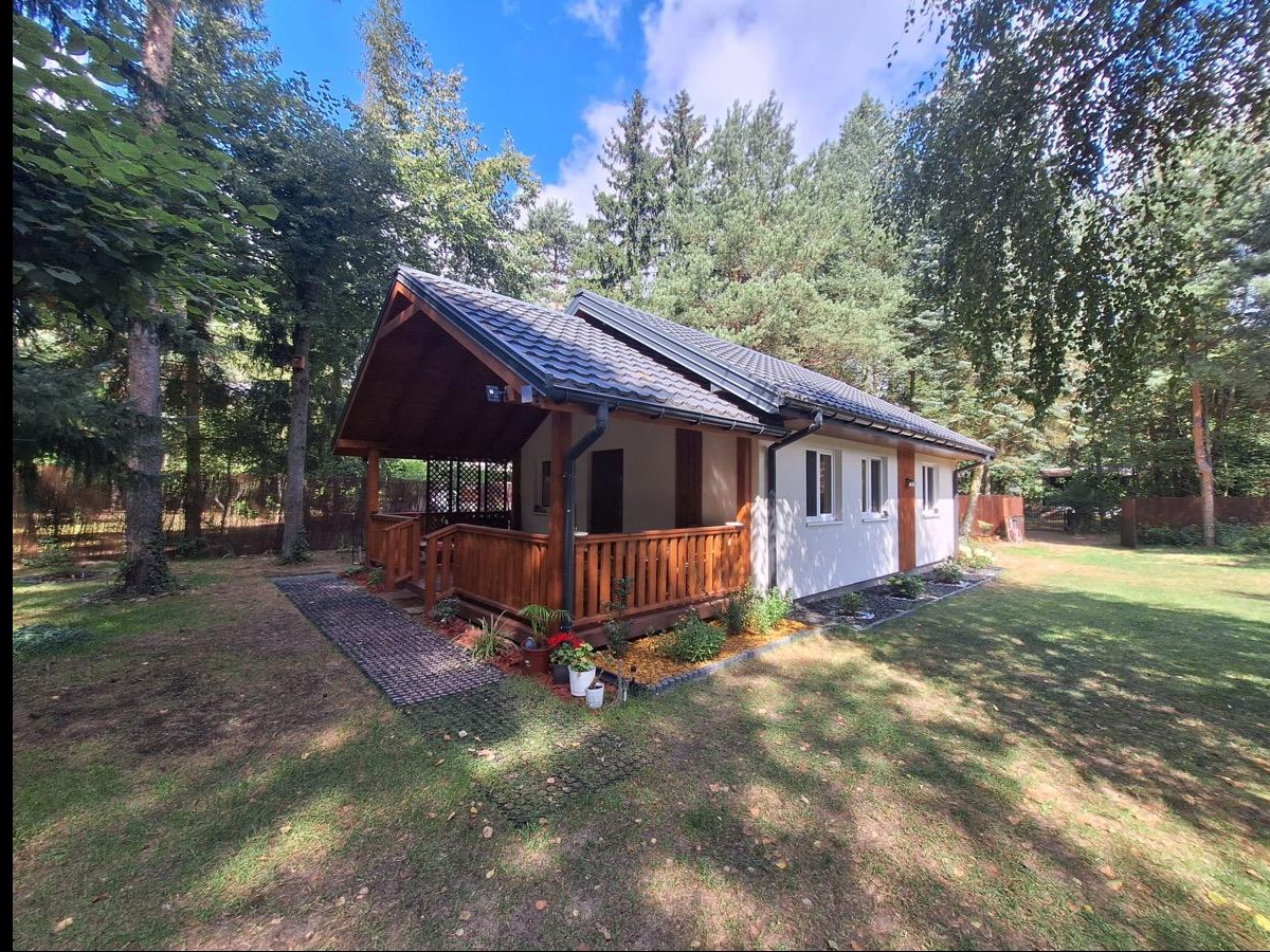 Parterowy dom z drewnianą werandą i szarą dachówką, otoczony zielenią. Widoczne kwiaty w donicach i ścieżka z ażurowych płyt. Ujęcie w słoneczny dzień.