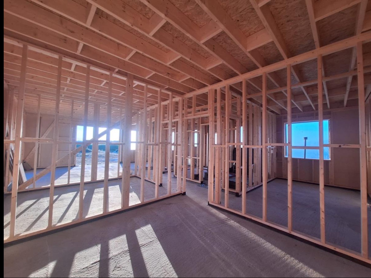 Wnętrze domu w trakcie budowy, widoczne drewniane ramy ścian i stropu, surowy beton na podłodze, wpadające światło przez okna. Konstrukcja w stanie surowym otwartym.