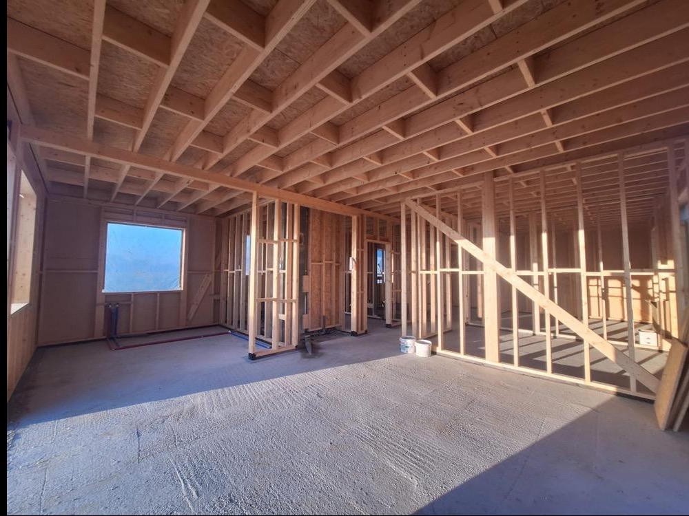 Wnętrze domu w trakcie budowy, widoczne drewniane ramy ścian i stropu, surowy beton na podłodze, okno z niebieskim tłem. Etap wznoszenia konstrukcji.