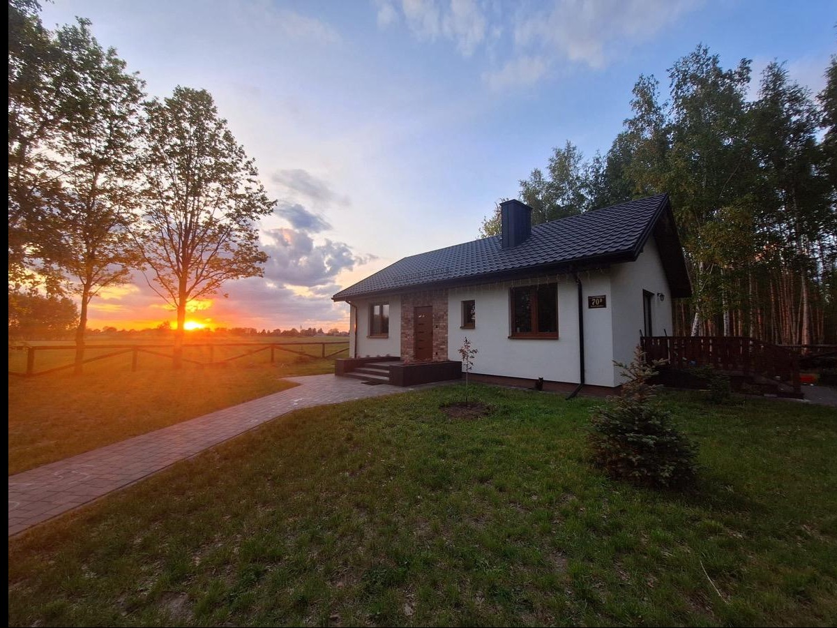 Parterowy dom z ciemnym dachem i jasną elewacją o zmierzchu. Widoczny ogród z ścieżką i las w tle. Na ścianie numer 70.