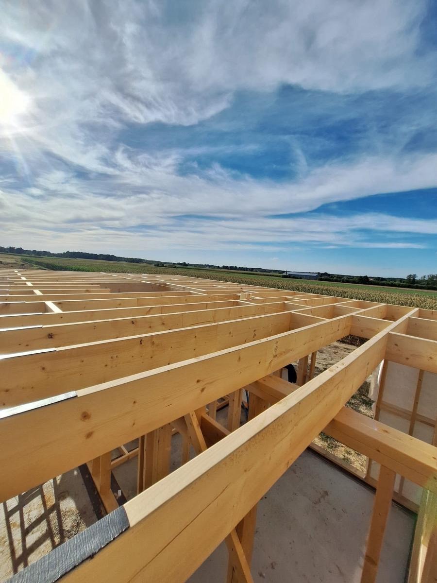 Wnętrze domu w trakcie budowy, widoczne drewniane belki konstrukcyjne na tle błękitnego nieba z chmurami i zielonego krajobrazu. Ujęcie z dołu, podkreślające surowość konstrukcji.