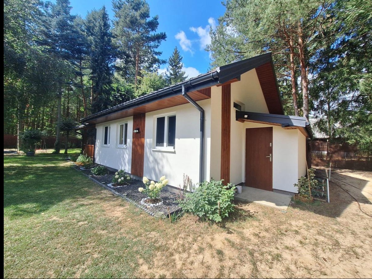 Parterowy dom z białymi ścianami i drewnianymi akcentami, otoczony zielenią. Widoczny fragment trawnika i drzewa w tle. Nowoczesna architektura w leśnym otoczeniu.