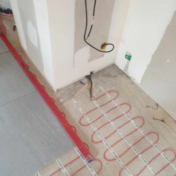 Taras Vysochynskyi - Montaż ogrzewania podłogowego: czerwone kable grzewcze ułożone na siatce, częściowo przykryte szarą płytką. Poziomica PRO przy ścianie. Widoczne kable elektryczne.