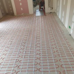 Taras Vysochynskyi - Rozłożona mata grzewcza z czerwonymi kablami na podłodze w trakcie prac wykończeniowych. Widoczne ściany przygotowane pod malowanie i fragment osoby w tle.