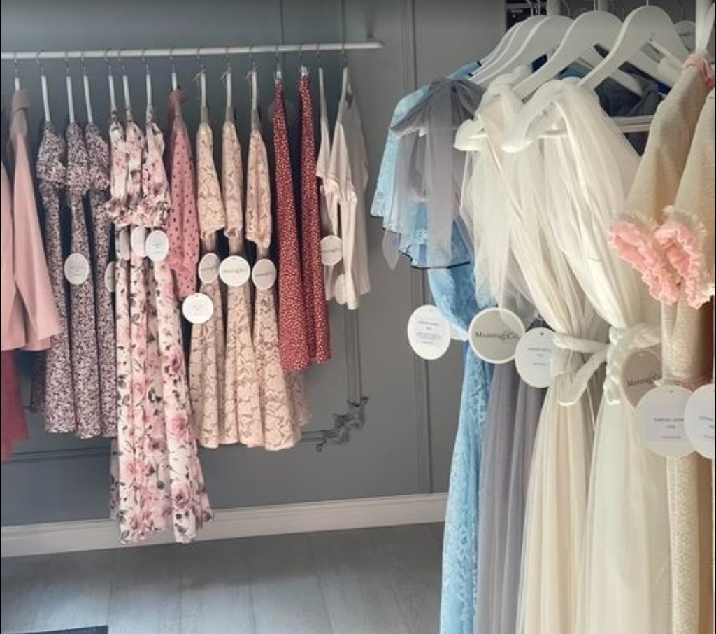 Różnorodne sukienki damskie, od zwiewnych, kwiecistych po eleganckie, tiulowe, wiszące na białych wieszakach w butiku. Widoczne metki z logo marki.
