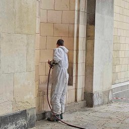 ADCLEANING M.LAMOWSKI K.JASIŃSKI S.C. - Pracownik w kombinezonie czyści kamienną elewację budynku za pomocą węża ciśnieniowego. Widoczne zabrudzenia i fragmenty oczyszczonej powierzchni.