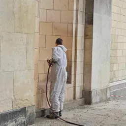 Pracownik w kombinezonie czyści kamienną elewację budynku za pomocą węża ciśnieniowego. Widoczne zabrudzenia i fragmenty oczyszczonej powierzchni.