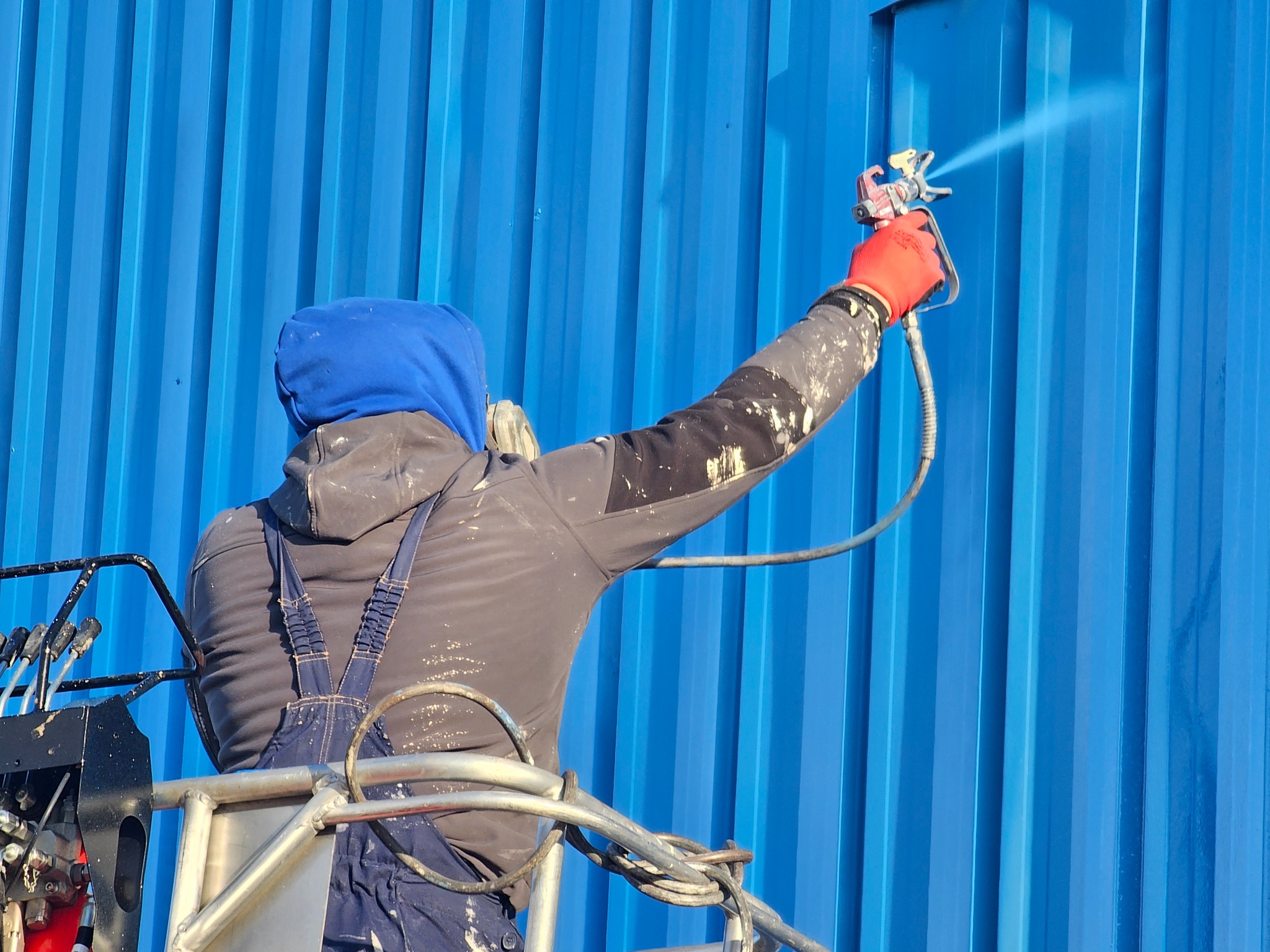 Pracownik w kombinezonie, z maską, maluje niebieską, metalową elewację natryskowo z podnośnika. Widoczny pistolet natryskowy i przewód doprowadzający farbę.