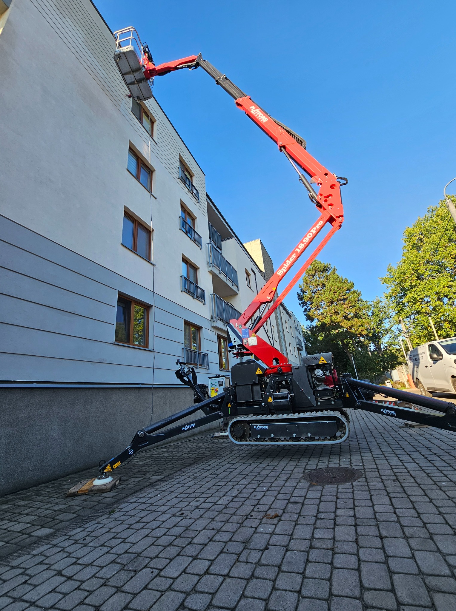 Czerwony podnośnik koszowy Spider 18.90 Pro przy białym budynku z balkonami. Widoczne zabezpieczenia pod stopami podnośnika. Błękitne niebo w tle.
