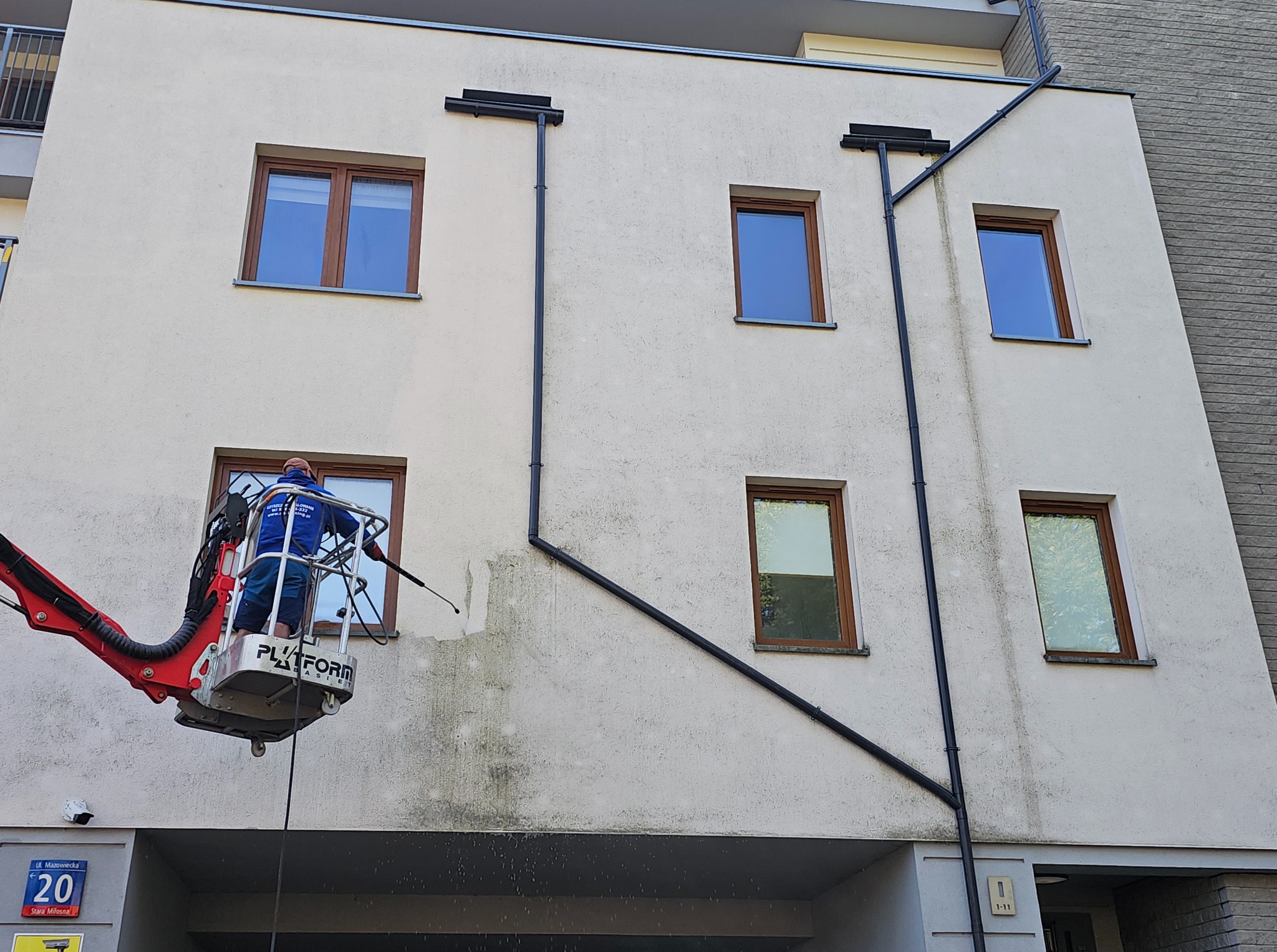 Pracownik na podnośniku koszowym myje elewację budynku. Widoczne różnice w czystości elewacji przed i po myciu. Ujęcie z dołu, fragment elewacji z oknami.