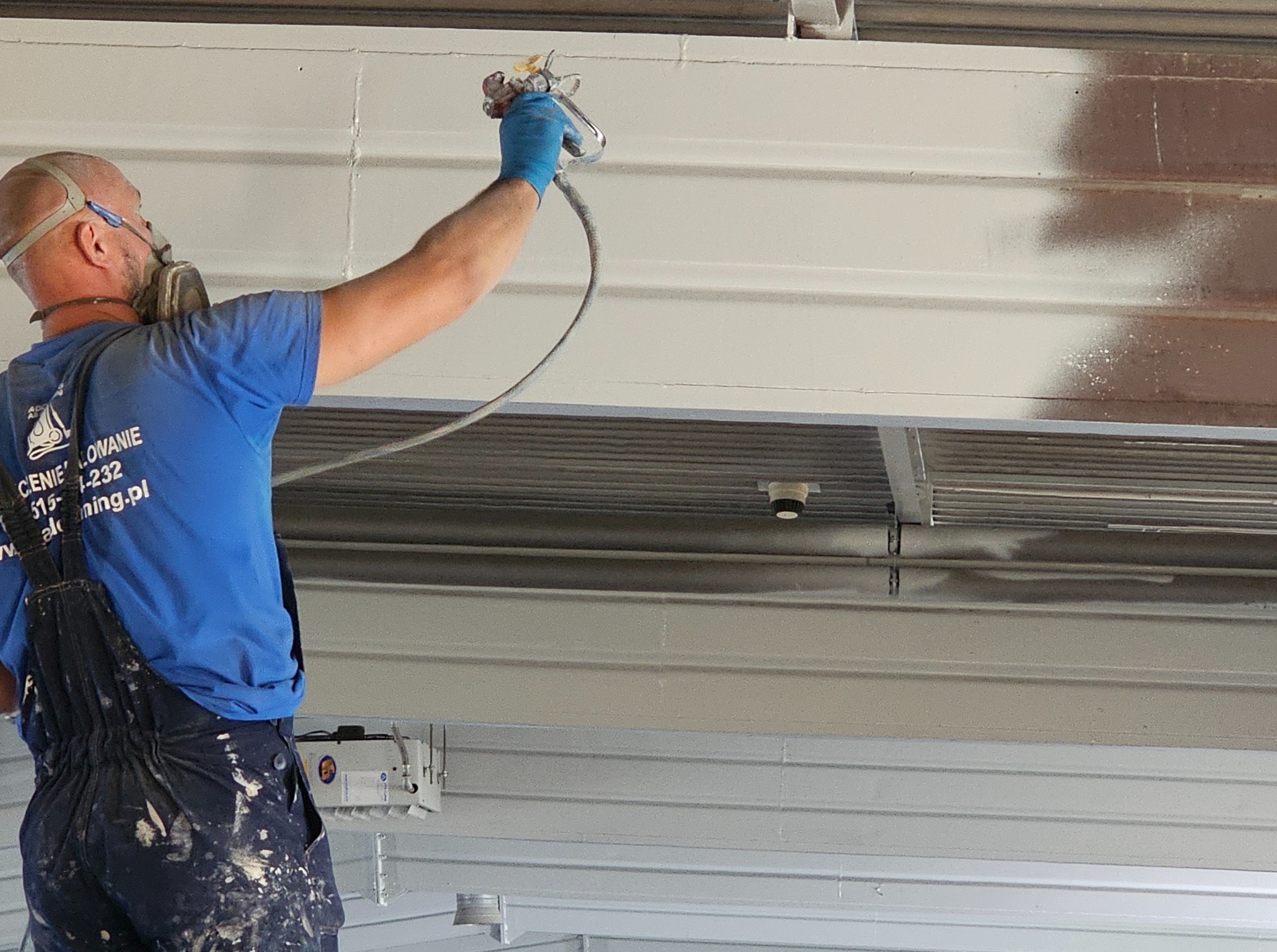Pracownik w masce maluje natryskowo metalową konstrukcję sufitu na biało. Widoczny fragment pomalowanej i niepomalowanej powierzchni. Ubranie robocze z plamami farby.