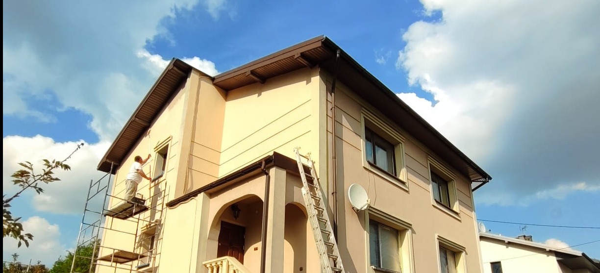 Pracownik na rusztowaniu maluje elewację domu w beżowym odcieniu. Widoczne okna z brązowymi ramami, brązowy dach i fragment błękitnego nieba z chmurami.