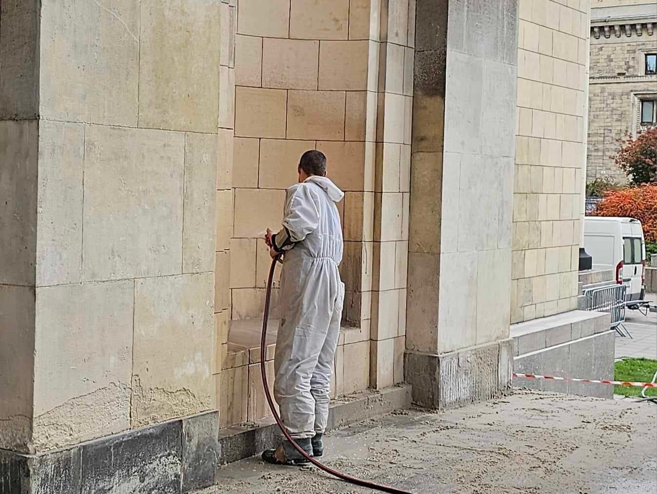 Pracownik w kombinezonie czyści kamienną elewację budynku za pomocą węża ciśnieniowego. Widoczne zabrudzenia i fragmenty oczyszczonej powierzchni.