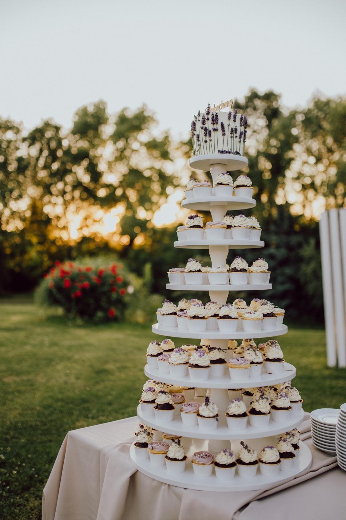 Wielopoziomowy stojak z babeczkami i tortem, dekorowany lawendą, na tle zielonego ogrodu z zachodzącym słońcem. Elegancki deser na przyjęcie.