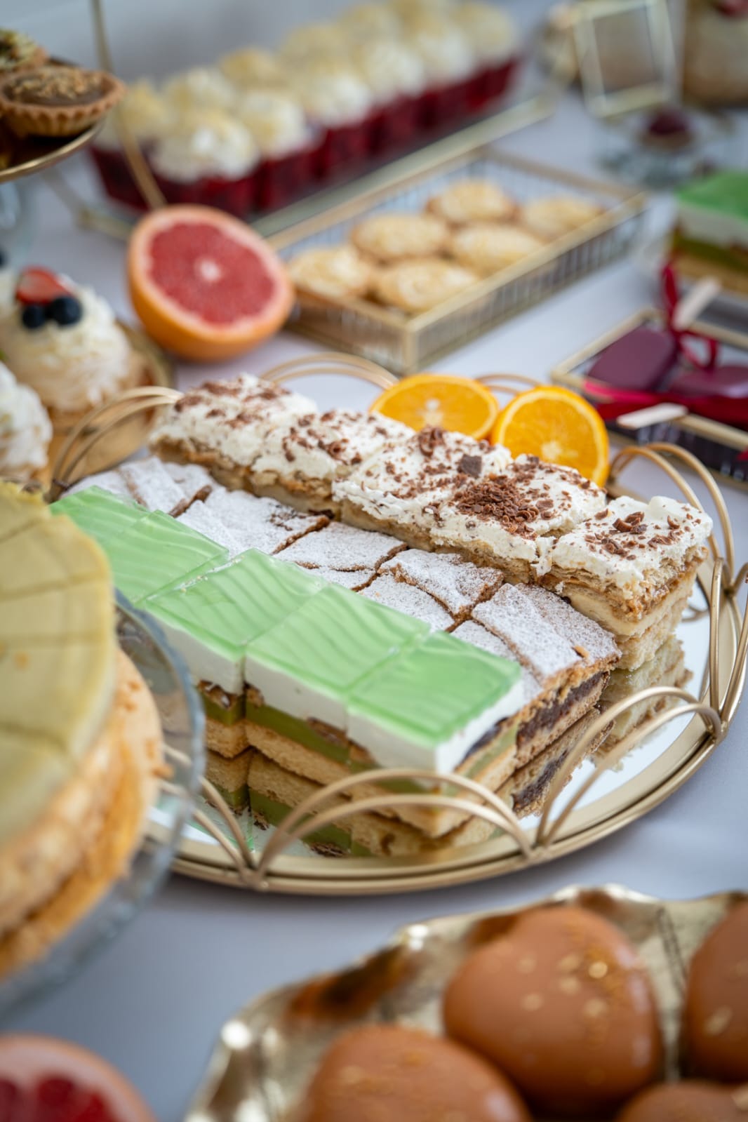 Elegancki catering: taca ze złotymi uchwytami, na niej ciasta z kremem, zieloną galaretką, posypane cukrem pudrem, obok pomarańcze i inne słodkości.