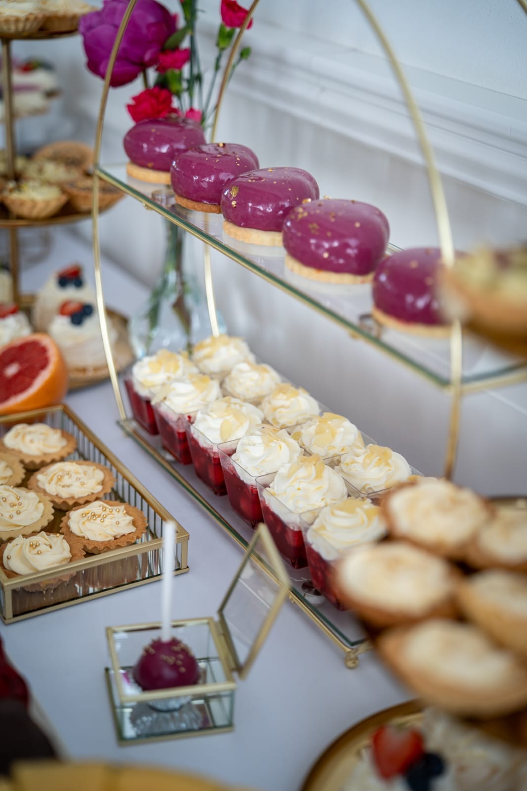 Elegancki stół z deserami: purpurowe monoporcje z jadalnym złotem, mini tartaletki z kremem, desery w kubeczkach z bitą śmietaną i owocami, całość na złotym stojaku.