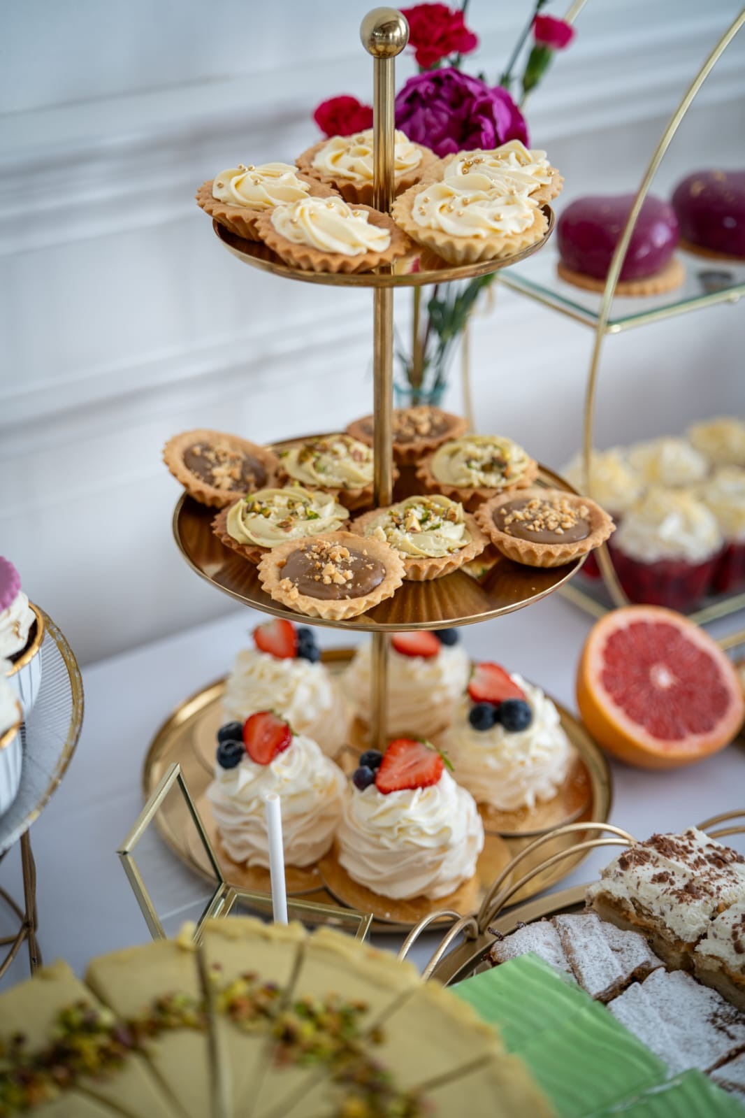 Elegancki, trzypoziomowy etażer z tartaletkami i bezami, dekorowany owocami i kwiatami. Widoczny grejpfrut i inne ciasta. Złote detale naczyń.