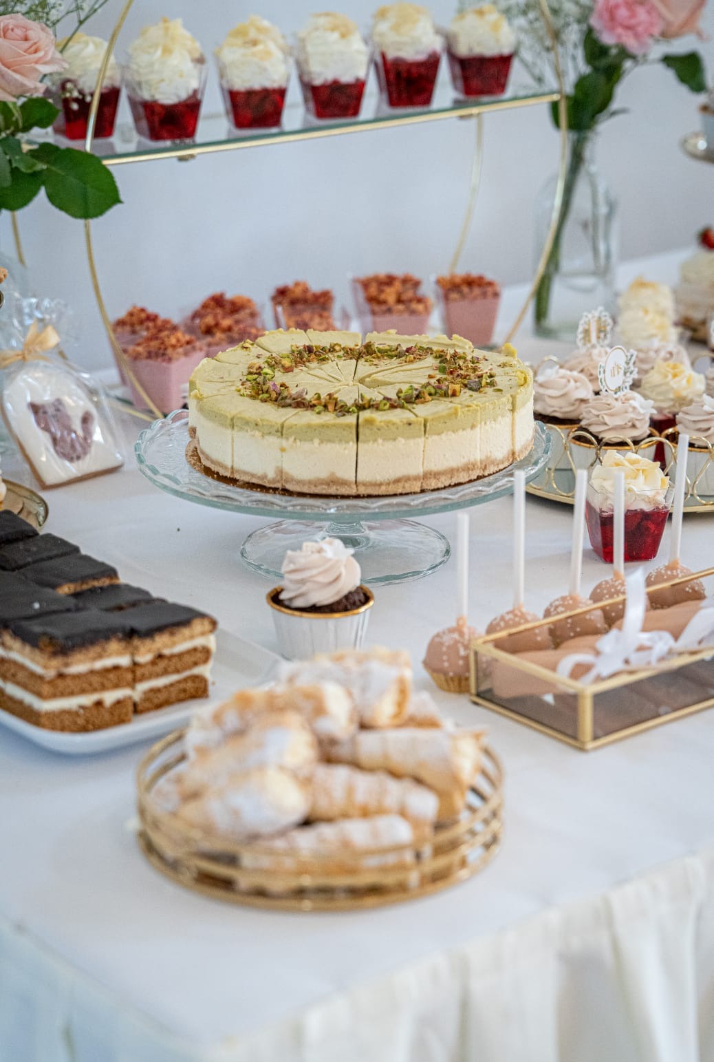 Elegancki stół z deserami: tort pistacjowy na szklanej paterze, babeczki, cake pops i kremowe desery w szklankach. Dekoracja kwiatowa i złote akcenty.