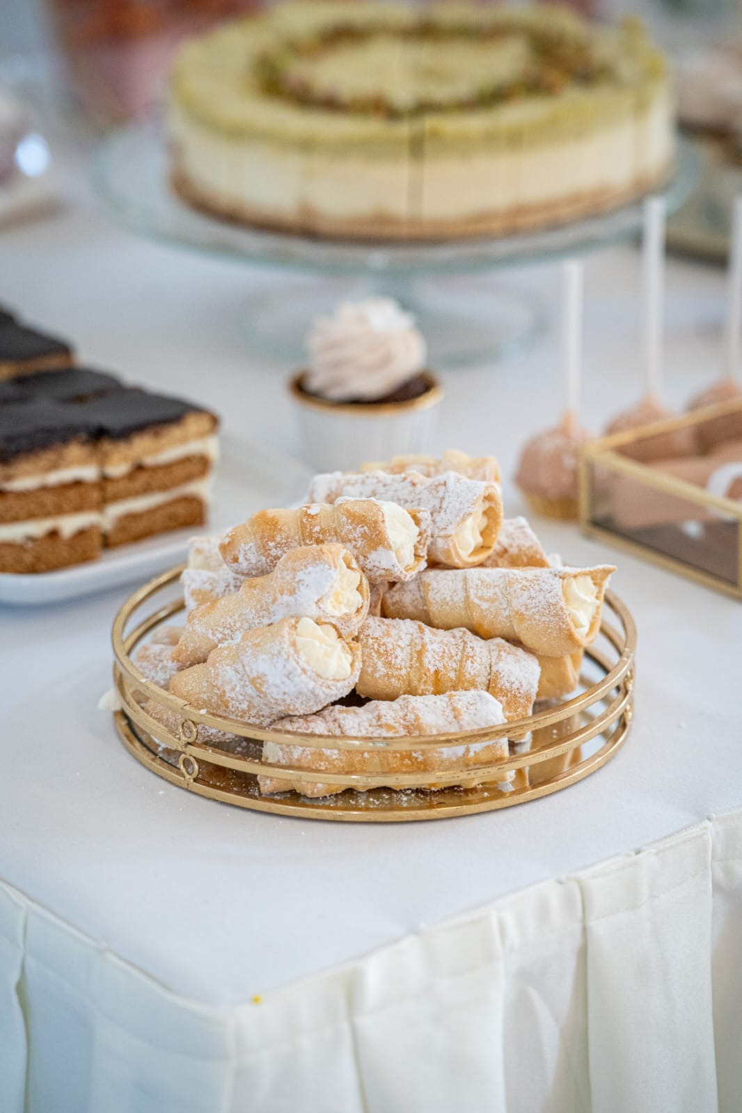 Złota patera z kremowymi rurkami oprószone cukrem pudrem, na stole z białym obrusem. W tle ciasta, babeczka i cake pops. Słodki stół na przyjęciu.