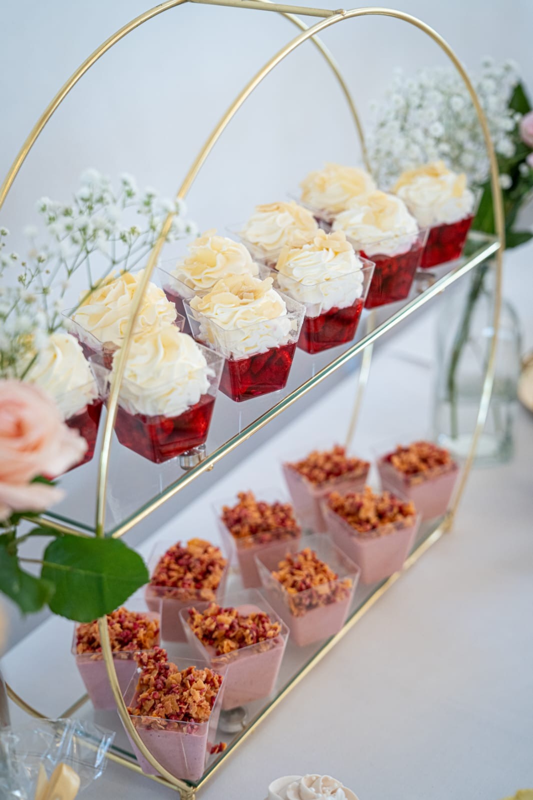 Deserowe porcje w przezroczystych pojemnikach na złotej, dwupoziomowej paterze. Na górze galaretka z bitą śmietaną i płatkami migdałów, na dole mus z posypką.