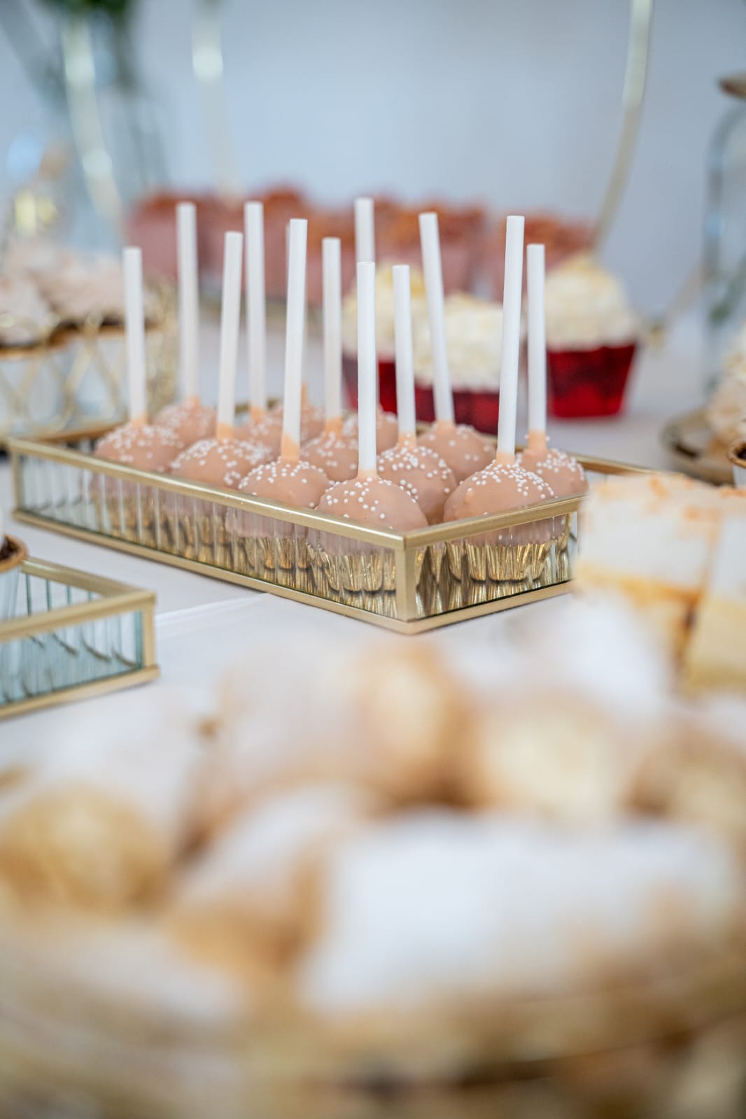 Beżowe cake pops z białymi posypkami w złotej, prostokątnej paterze. W tle inne desery i dekoracje na stole. Słodki stół na przyjęciu.