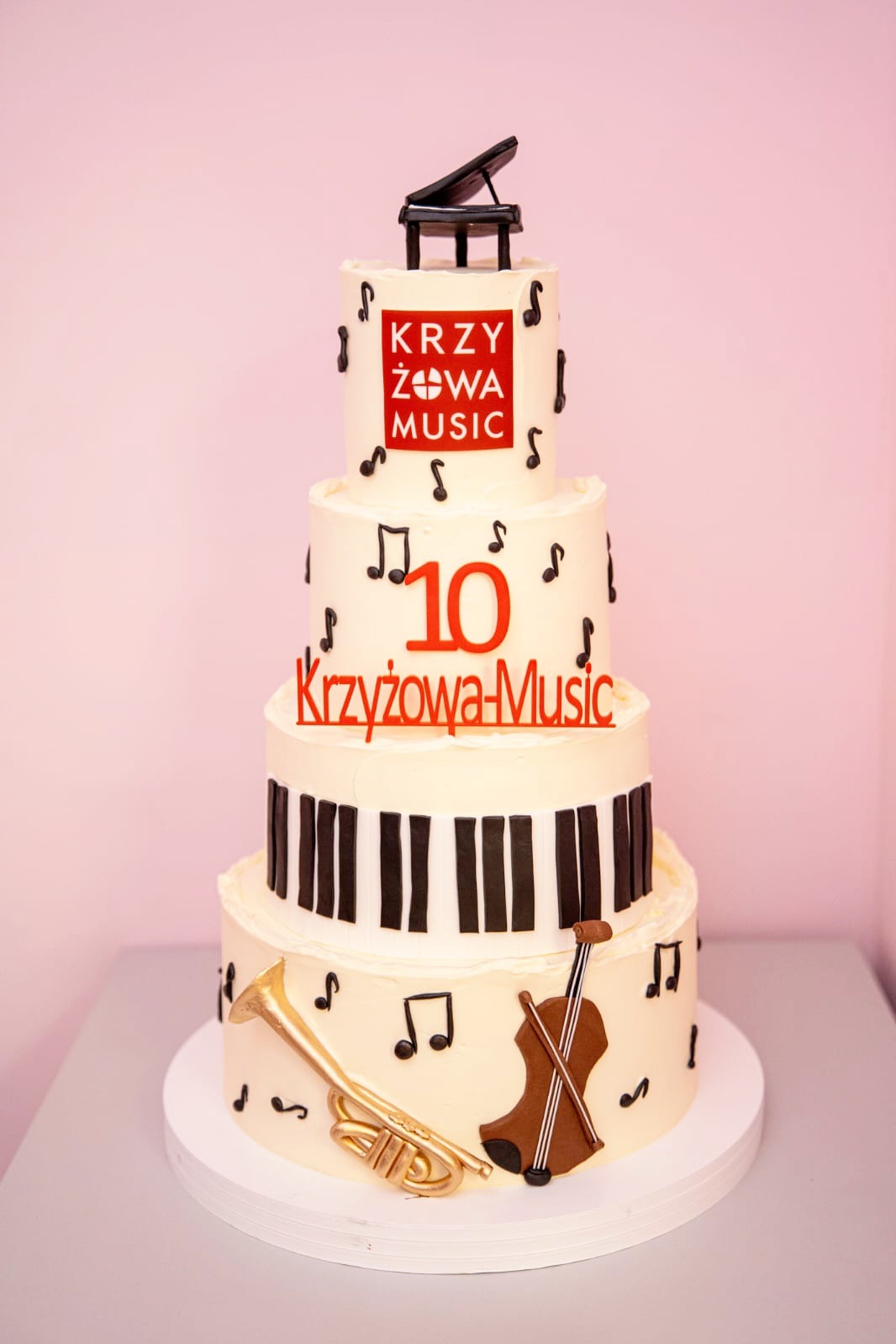 Wielopoziomowy tort z motywami muzycznymi: pianino, klawiatura, nuty, trąbka i skrzypce. Dekoracja z logo 'Krzyżowa Music' i cyfrą 10. Kremowe tło, jasne oświetlenie.
