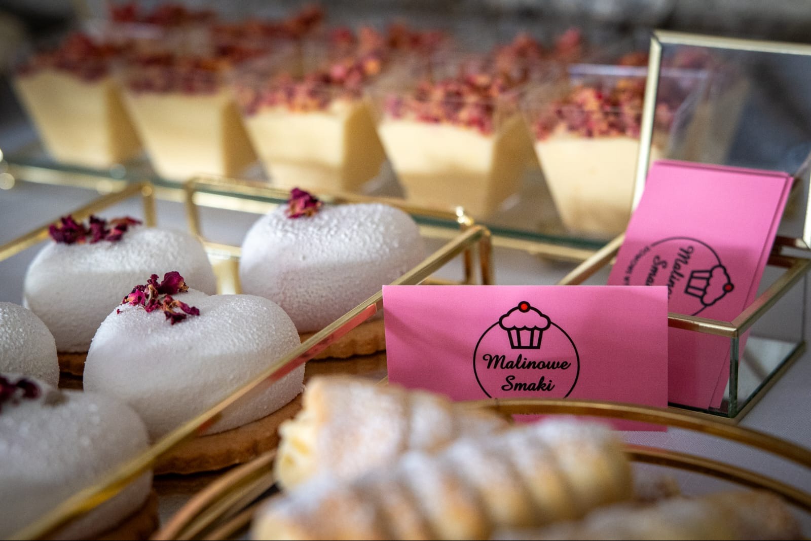 Eleganckie desery: białe serca z różanymi płatkami, kremowe kostki i różowe wizytówki 'Malinowe Smaki' na złotych paterach. Słodki stół w zbliżeniu.