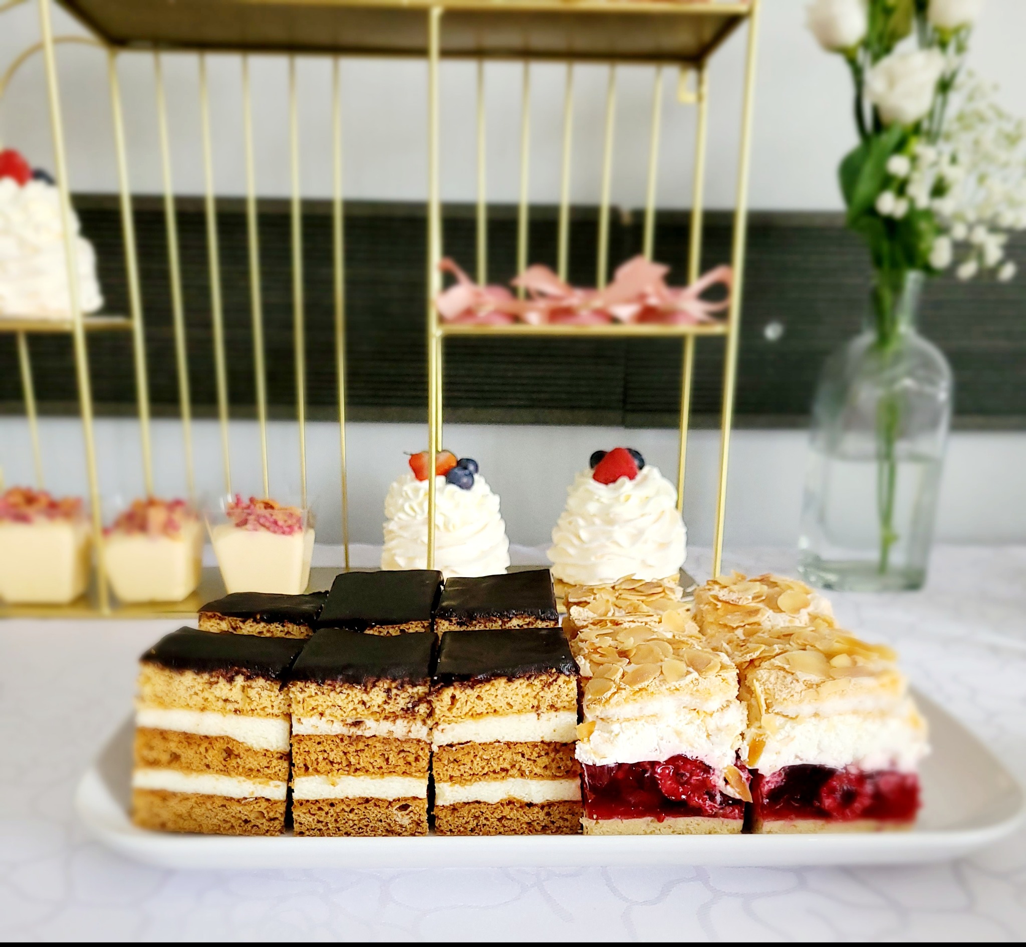 Elegancki stół z deserami: kremowe babeczki z owocami, ciasto miodowe z czekoladą i tort z wiśniami, całość na białym talerzu, w tle złota dekoracja i kwiaty.