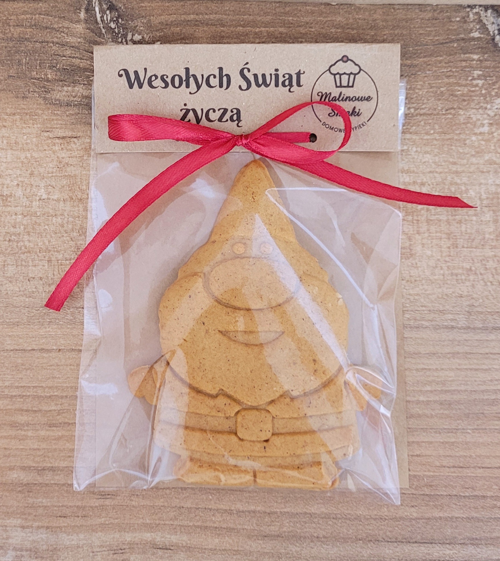 Piernikowy skrzat w przezroczystym opakowaniu z czerwoną wstążką i etykietą 'Wesołych Świąt' na drewnianym tle. Domowe wypieki.
