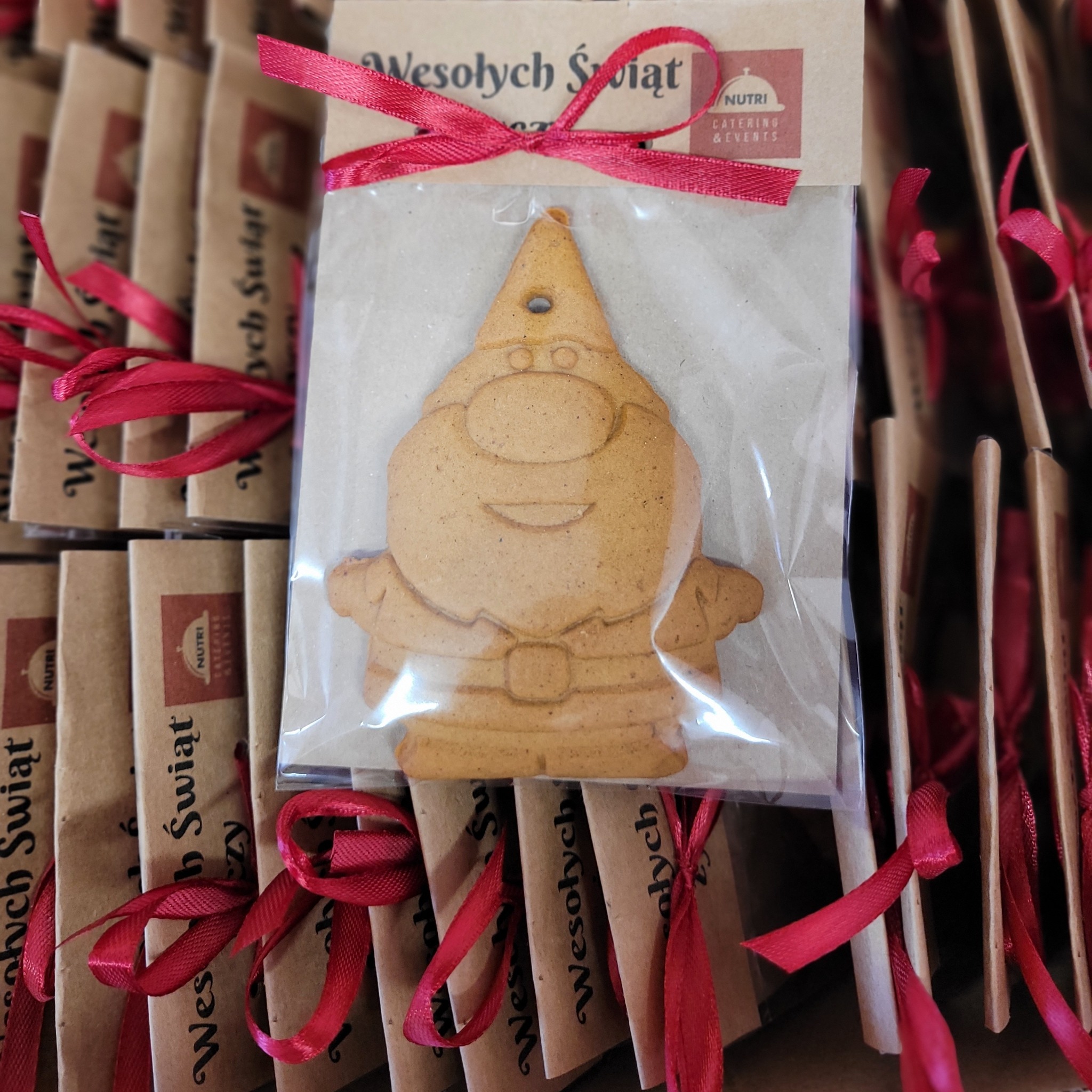 Piernikowy skrzat w folii z czerwoną wstążką i napisem 'Wesołych Świąt', część świątecznego zestawu od Nutri Catering & Events. Ułożone jeden za drugim.