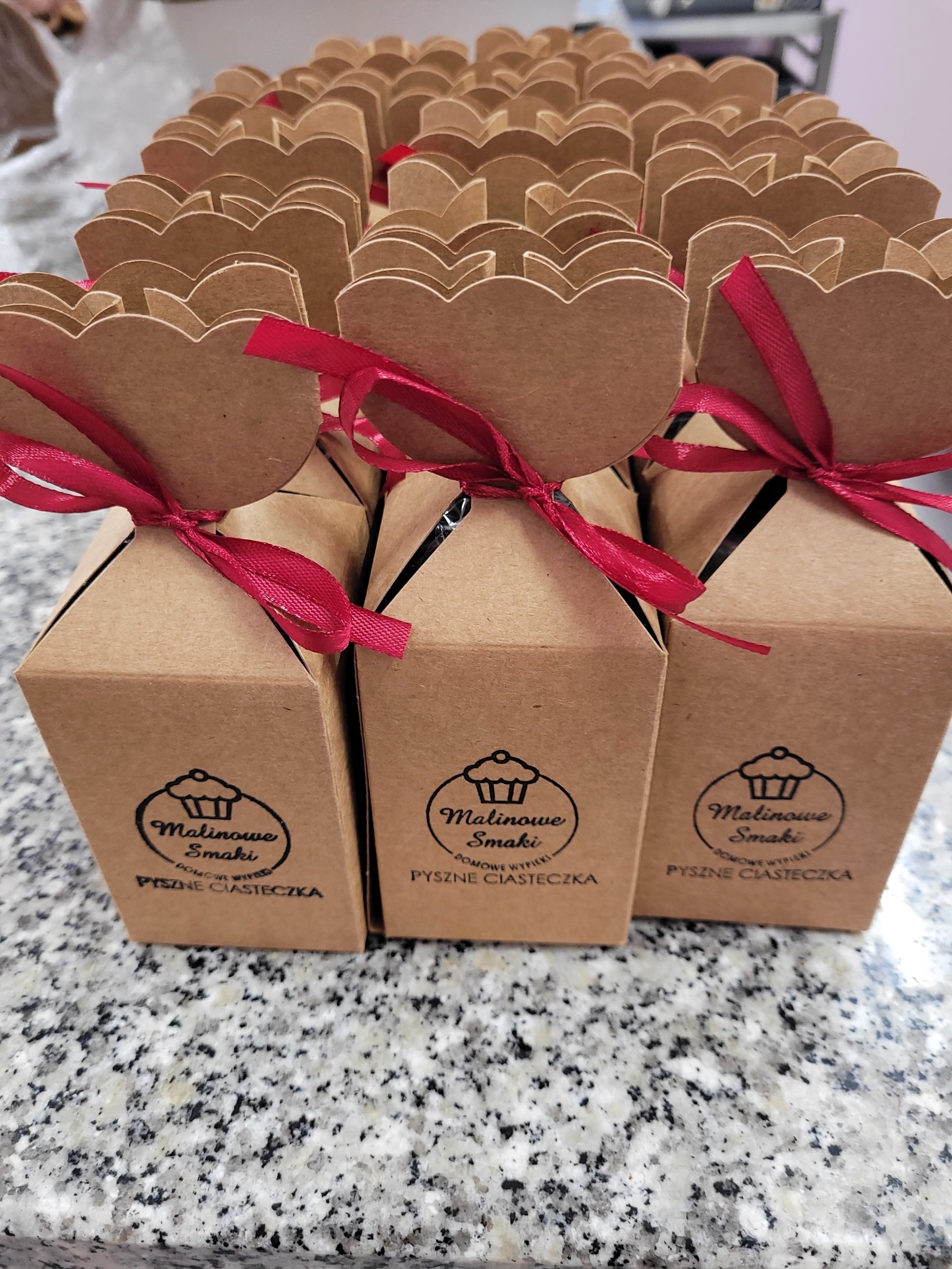 Rząd pudełek z ciastkami 'Malinowe Smaki' z dekoracją w kształcie serca i czerwoną wstążką, ustawionych na blacie z granitu. Widoczne logo firmy na pudełkach.